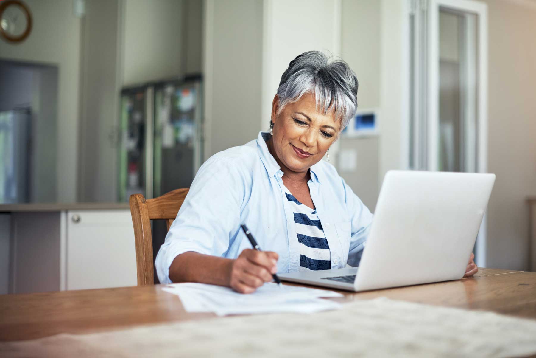 senior woman taking notes while sitting at her kitchen table in front of her laptop